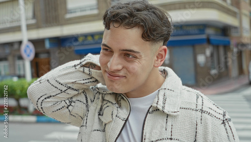 Young hispanic man with hand on neck wearing white check jacket on urban street beside storefront, crosswalk and traffic sign; thoughtful shy.