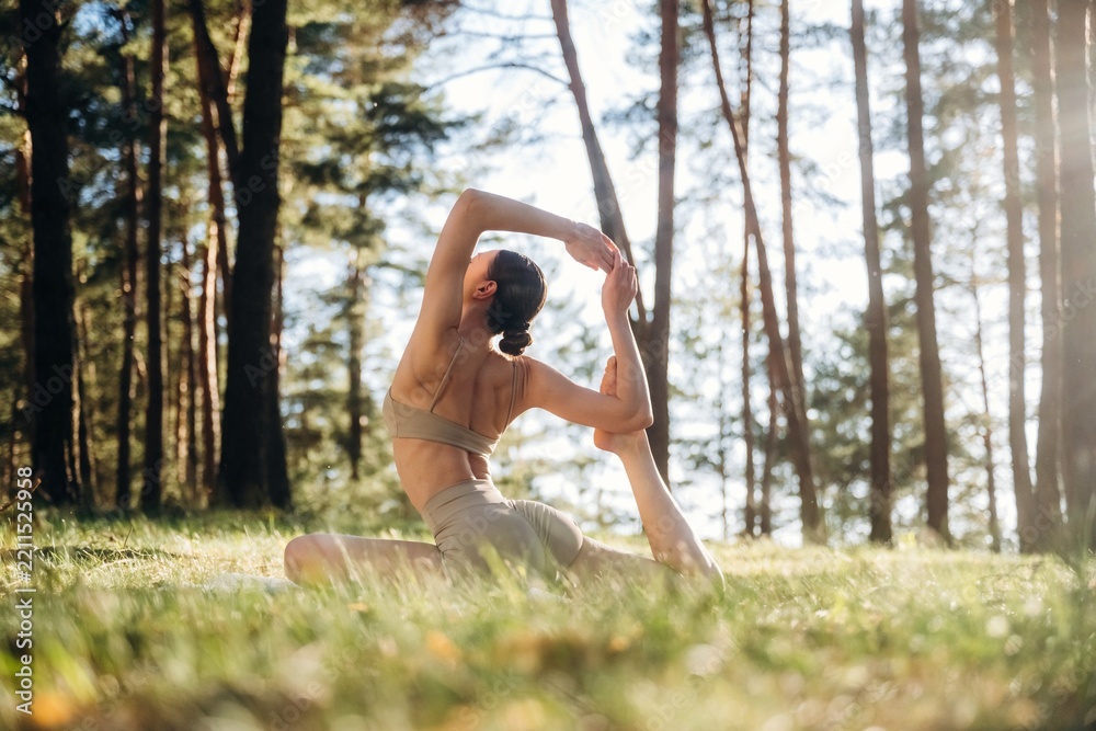 Obraz premium Female yoga practitioner in a forest performs a flexible pose on grass, surrounded by tall trees and dappled sunlight filtering through the foliage