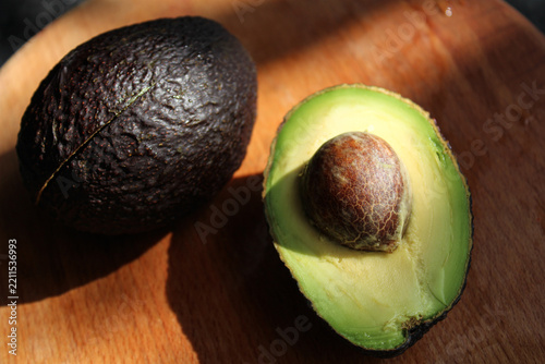 Fresh ripe Hass avocado halved with seed and a whole fruit on wooden cutting board in bright natural sunlight with deep shadows