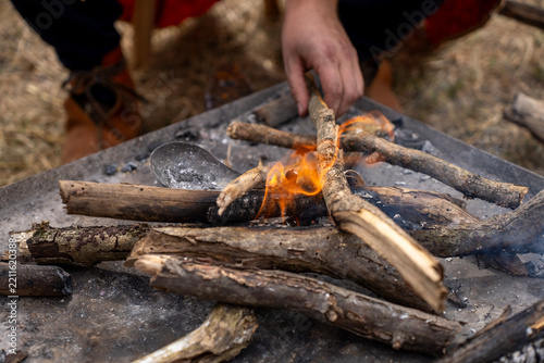 A hand places a branch on a small outdoor fire among logs, flames, and ash.