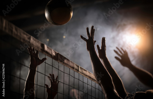 Volleyball players reaching for the ball during a match in a dimly lit gymnasium