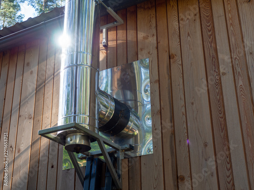 Stainless steel chimney and flue pipe installed on the exterior wall of a wooden building, with reflective heat shield and metal support structure. 