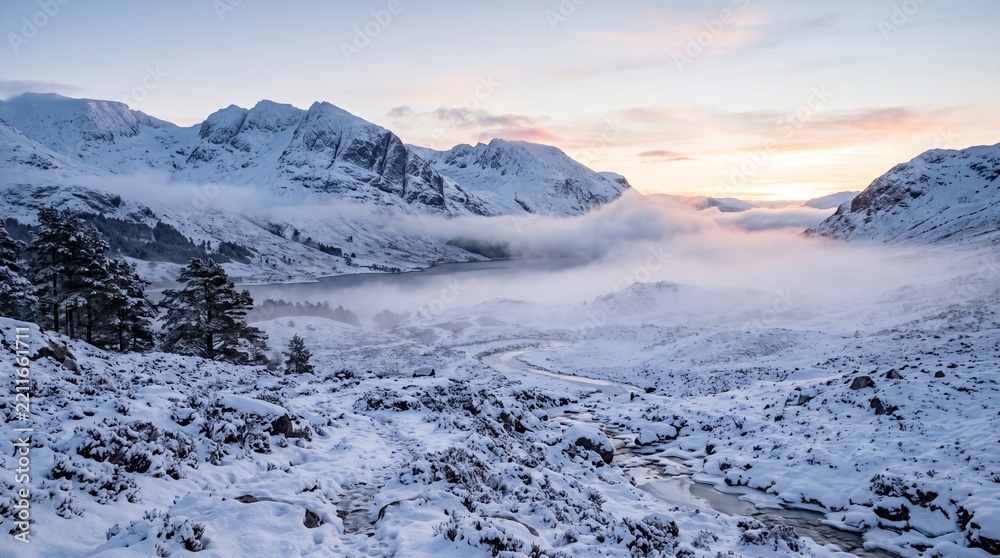 Obraz premium Snow covered Scottish Highlands landscape at sunrise with mist rolling over a loch and mountains