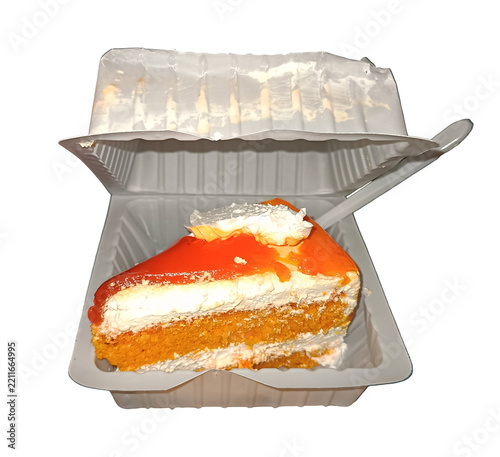 A delicious slice of orange cake with cream frosting, presented in a disposable plastic clamshell container with a plastic spoon, isolated on white.