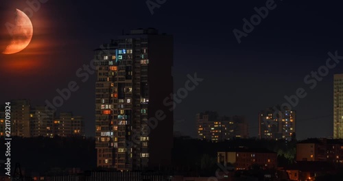 Beautiful dramatic time lapse of a large orange crescent moon setting behind a modern city residential building at night