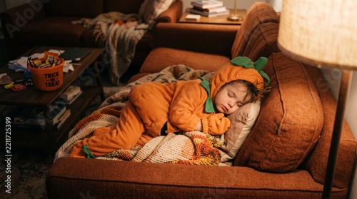 Adorable toddler dressed in a pumpkin costume sleeping soundly on a cozy couch after trick-or-treating