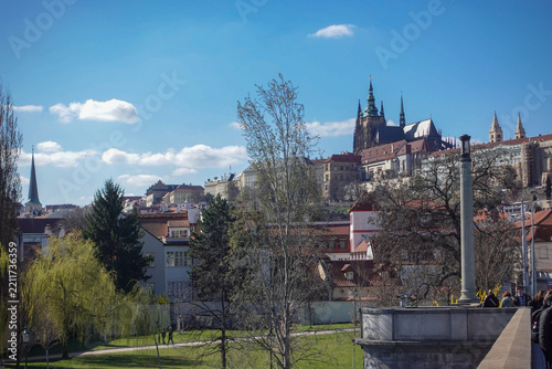 Panoramic cityscape of Prague displaying red terracotta rooftops, historic spires, and the Vltava River spanning across the urban horizon.