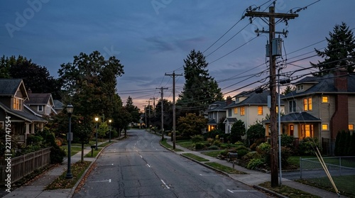Suburban street at dusk, residential neighborhood with streetlights and overhead power lines.