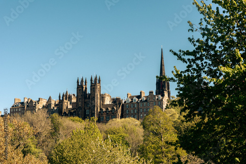 Daylight horizontal photograph showing the historic stone buildings and Gothic spire of Edinburgh's Old Town in Scotland, framed by trees under a clear blue sky