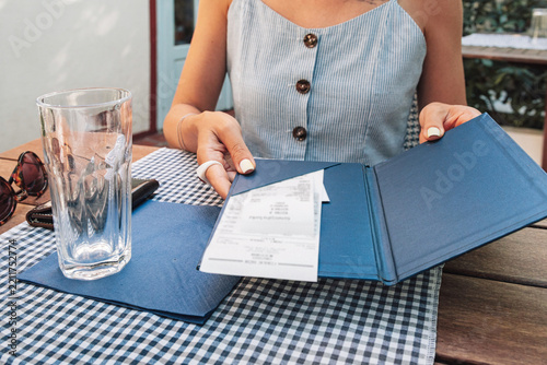 Woman's hands holding a blue folder with a paper receipt, preparing to pay for a meal at an outdoor restaurant