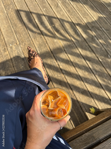 Woman hand holding iced coffee drink on sunny outdoor wooden terrace with leopard print shoes for fashion lifestyle and coffee break concept