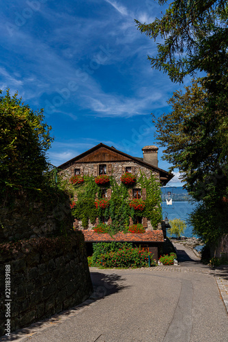A rustic stone house draped in ivy and red flowers stands at the end of a shaded lane in Yvoire, opening onto a glimpse of Lake Geneva beyond.