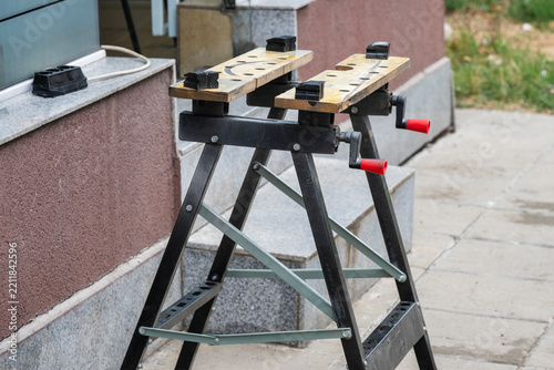 A portable folding workbench with built-in clamping jaws and crank handles stands outdoors on a paved patio beside house steps