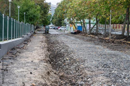 A torn-up urban street and excavated sidewalk trench filled with loose crushed gravel run parallel to a green metal fence and tree line during municipal roadbed reconstruction works.