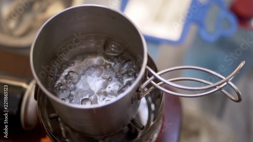Water vigorously boils inside a stainless steel metal cup over a portable gas burner. Bubbles rapidly churn across the heated liquid surface while metal wire handles extend outward.