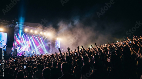 Large crowd of people with hands raised enjoying a live music concert at night with bright stage lights and smoke effects