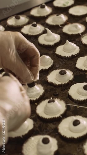 Close up video of a pastry chef in white gloves meticulously placing chocolate drops on freshly piped white meringue swirls on a chocolate base. A tray full of treats in a bakery