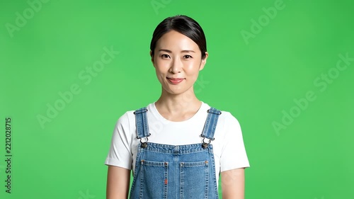 Young Asian woman in denim overalls smiling against a green screen background