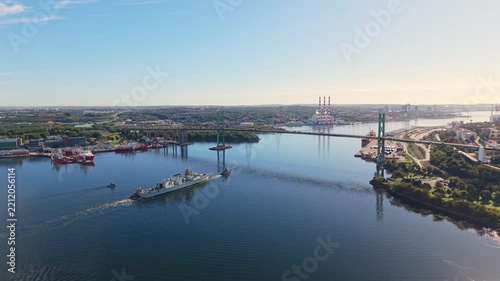 Aerial view of a military towing operation in Halifax Harbour, Nova Scotia, with a military ship pulling another vessel beneath a bridge while creating long wakes across the calm morning water.