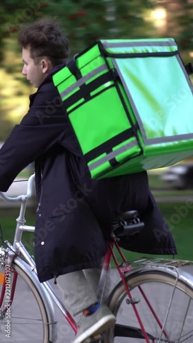 Young courier rides a bicycle with a large bright green insulated food delivery backpack in an urban park.