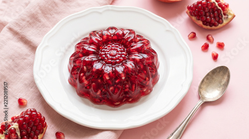 Ruby red pomegranate gelatin dessert on a scalloped ceramic plate with fresh seeds and silver spoon arranged on a soft pink table for festive dining fruit recipes and holiday celebrations