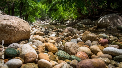 Smooth unpolished stones fill a shallow riverbed with lush green foliage bordering the clear flowing water