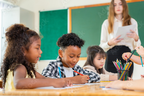 Multiethnic school children drawing with colored pencils in classroom with teacher, diverse students learning art and writing at desk with educator supervision