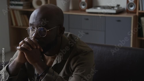 Middle aged African American man in glasses sitting with clasped hands experiencing stress and emotional burnout indoors. Clip ideal for mental health awareness and therapy campaigns