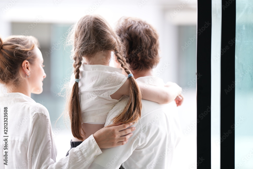 Fototapeta premium Happy young family with little daughter appear at the glass facade holding their daughter standing on the terrace of a newly built suburban home. Happy family lifestyle and wellbeing concept