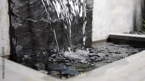 Gentle torrent of crystal-clear water pours over a textured stone surface, creating soothing sounds and a calming atmosphere within a beautifully designed outdoor space surrounded by greenery.