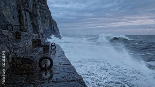 Dynamic waves collide with natural stone walls, creating a mesmerizing display of white foam and crashing sounds, all under the soft glow of twilight's sky.