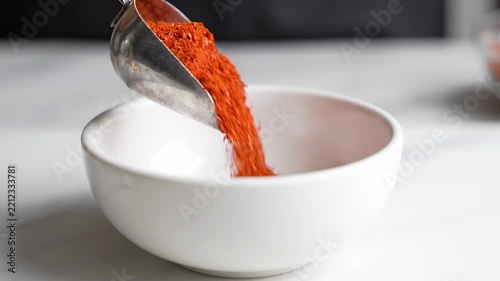 Bright red spice cascades into a smooth white bowl, illustrating a vibrant kitchen moment. The hand's gentle motion highlights the artistry of cooking during a sunny afternoon.