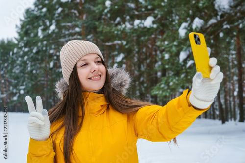 Winter selfie in snowy forest happy woman in yellow jacket showing peace sign while taking photo on smartphone