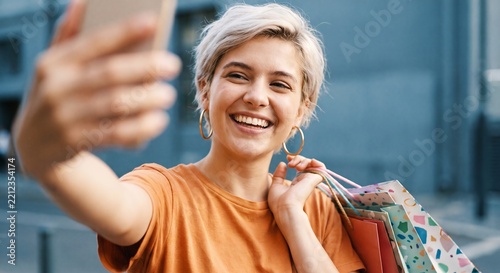 Happy young woman taking a selfie outdoors while holding colorful shopping bags and smiling cheerfully during a retail therapy day in the city