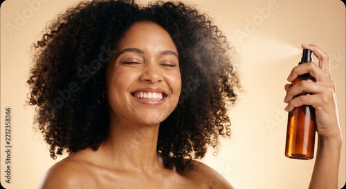 Smiling young woman with curly hair spraying refreshing facial toner from a bottle during her daily beauty skincare routine