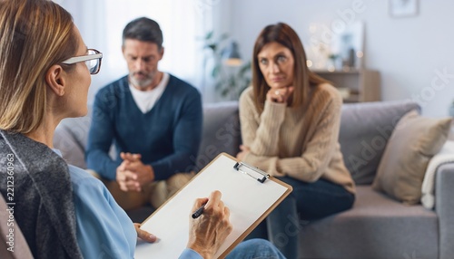A professional psychologist taking notes during therapy session with an unhappy middle aged couple sitting on couch discussing relationship issues and marriage crisis