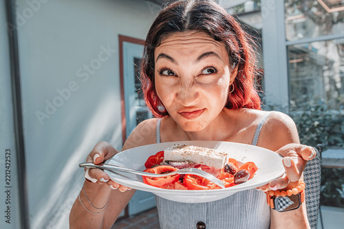 Playful reaction shot for food blogs and restaurant: woman making funny face while holding Greek salad plate with feta. Dining concept.