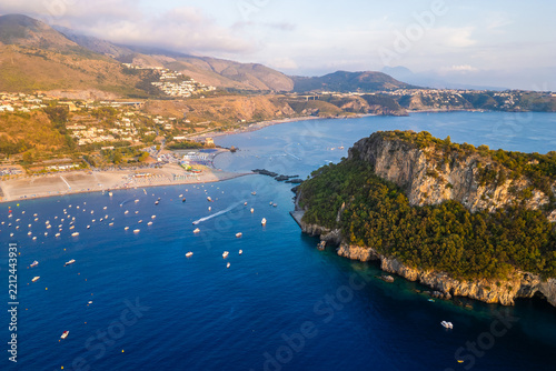 A breathtaking sunset aerial view captures Isola del Dino and the bay at Praia a Mare in Calabria, Italy. Boats navigate the deep blue waters along a sprawling beach, framed by dramatic coastal