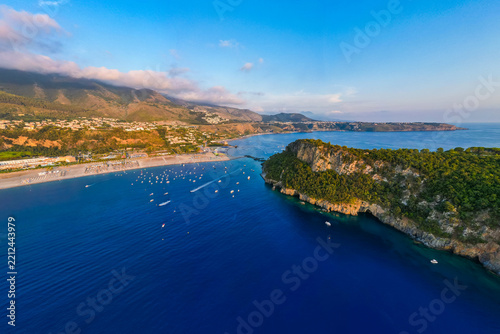 A breathtaking sunset aerial view captures Isola del Dino and the bay at Praia a Mare in Calabria, Italy. Boats navigate the deep blue waters along a sprawling beach, framed by dramatic coastal