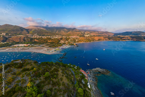 A breathtaking sunset aerial view captures Isola del Dino and the bay at Praia a Mare in Calabria, Italy. Boats navigate the deep blue waters along a sprawling beach, framed by dramatic coastal
