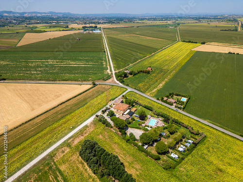 An aerial drone view reveals a charming Tuscan agriturismo nestled among vibrant sunflower fields and vast farmland. The property features a swimming pool and an organized camperstop for motorhomes in