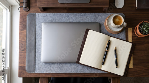 top down flat lay of elegant wooden work desk featuring grey felt organizer pad closed laptop computer open journal notebook fountain pens cup of coffee wooden ruler and green succulent