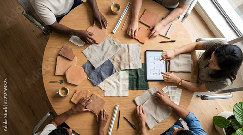 Overhead view of multi-ethnic creative interior design team seated around circular wooden table reviewing terracotta tiles fabric swatches and architectural floor plan on digital tablet