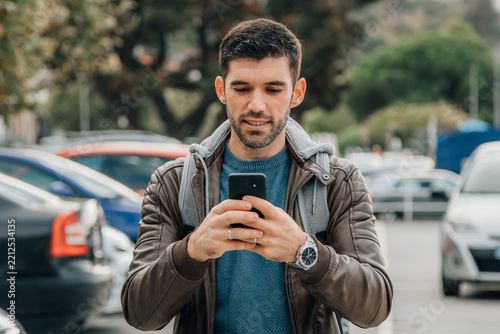 Young man with beard smiling while checking smartphone on urban street with parked cars in background