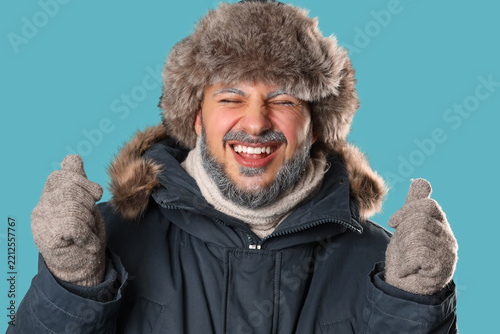 Young man in warm gloves, winter clothes and with frosted beard on blue backg...