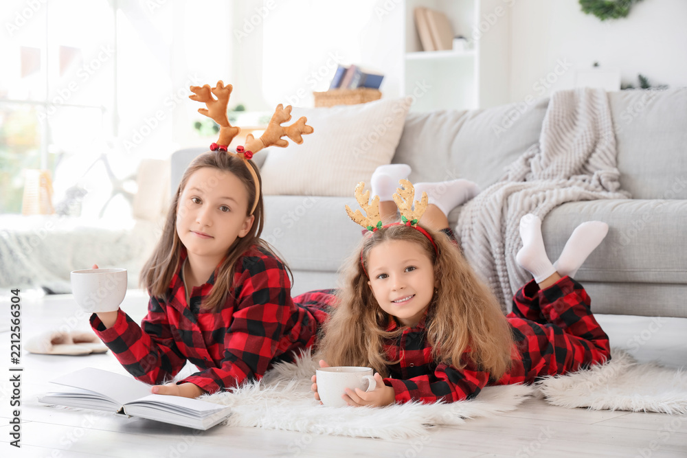 Obraz premium Cute sisters in reindeer horns with cocoa reading story at home on winter day