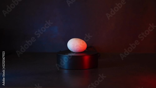 Single white egg rotating 360 degrees on a sleek black pedestal. Soft red and blue lighting creates a mysterious minimalist studio scene with a dark background.