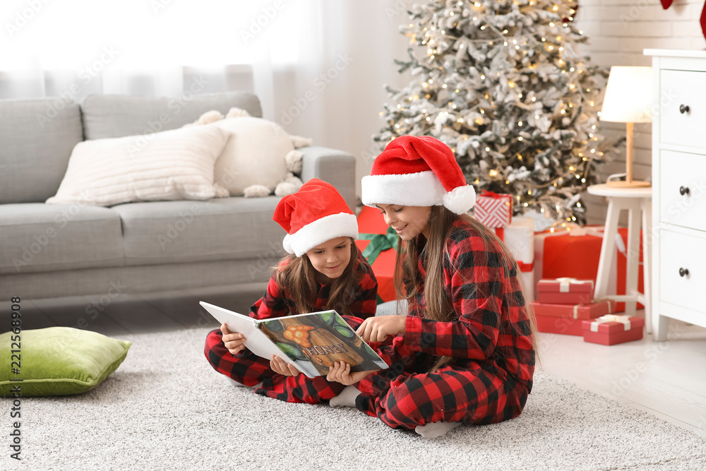 Obraz premium Cute happy little girls in checkered pajamas and Santa hats with book reading Christmas story at home