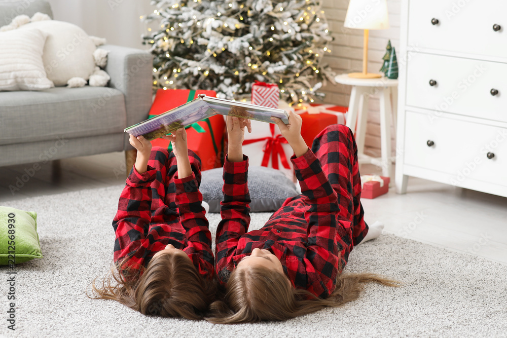 Obraz premium Cute little girls in checkered pajamas with book reading Christmas story at home
