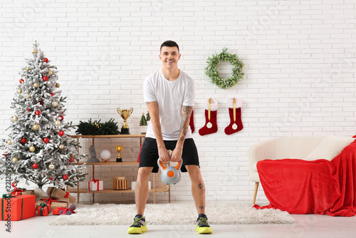Young happy man training with kettlebell in living room decorated for Christmas